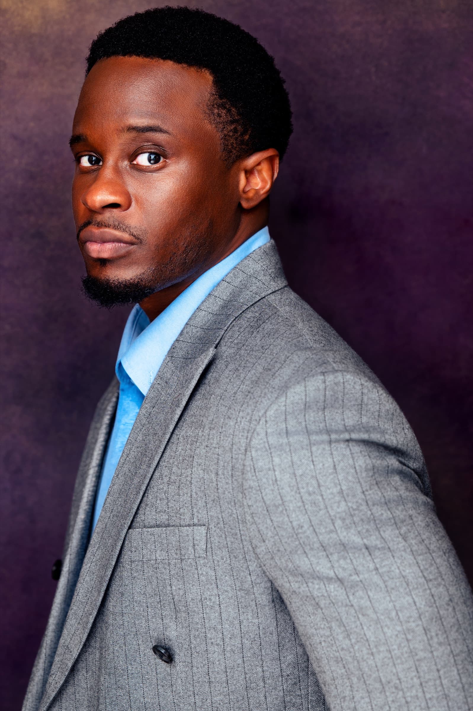 Lanre Adigun headshot — grey pinstripe suit, purple backdrop